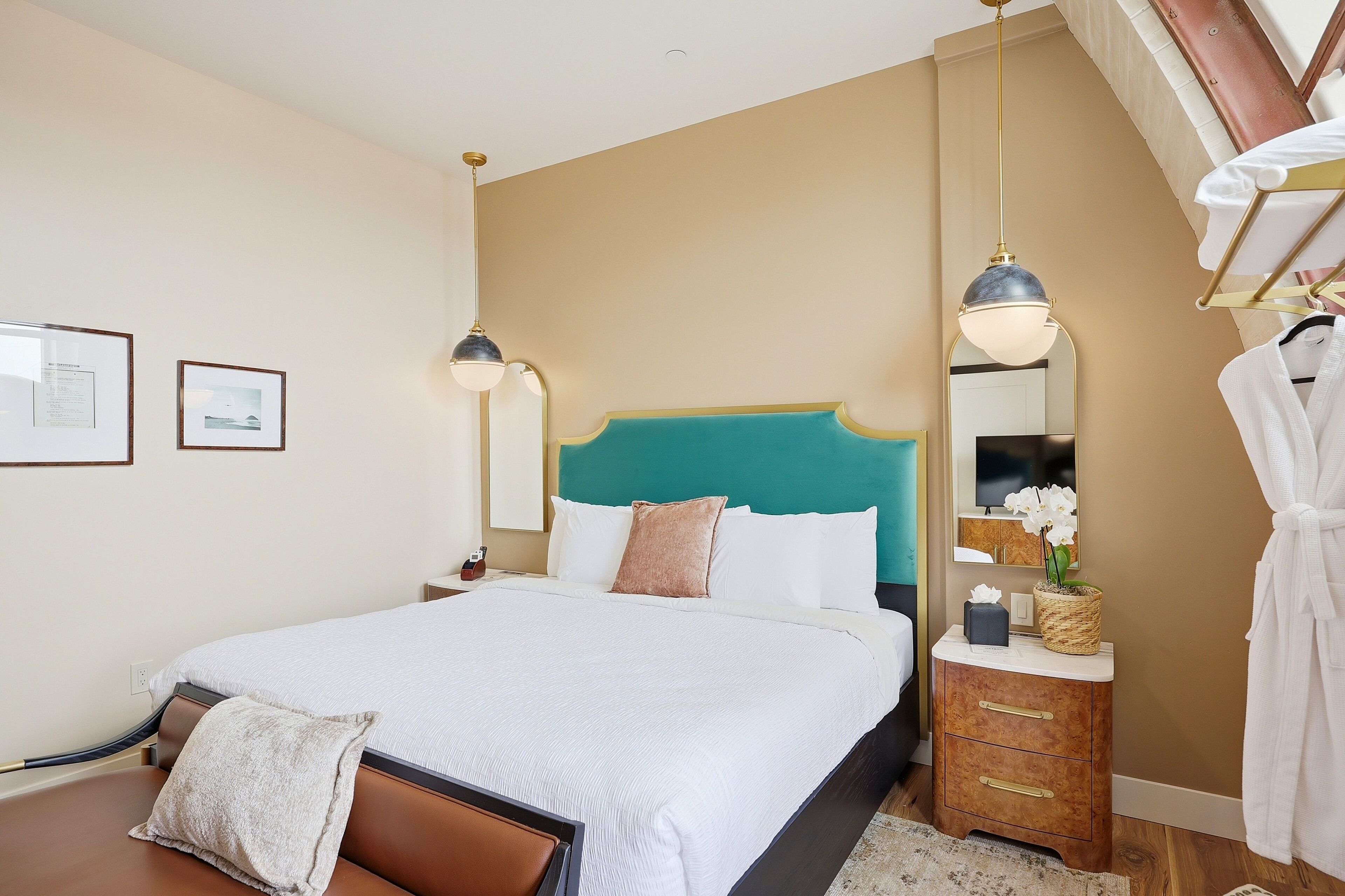 Una cama bien hecha con un cabecero verde aguamarina, una mesita de noche con una lámpara y un espejo con una orquídea en maceta.
