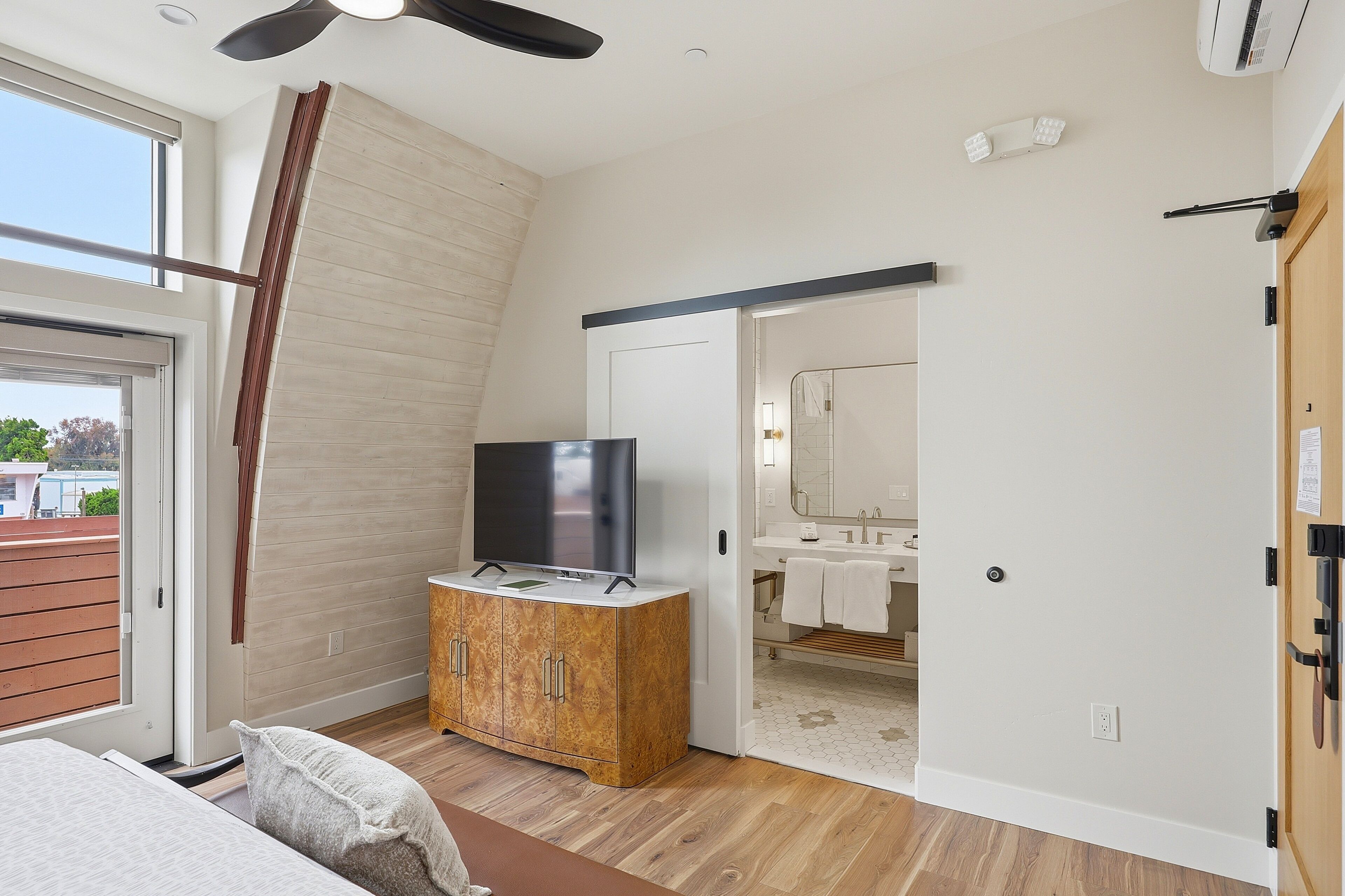 A bedroom with a TV on a wooden cabinet, a sliding door to a bathroom, and a view of the outside.