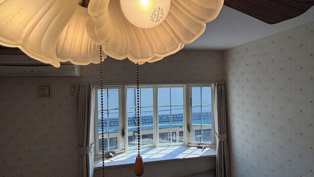 A room with a ceiling fan, two beds, a window with curtains, and a view of the cityscape.