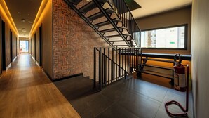 Um corredor de hotel moderno com piso de madeira, parede com padrão de tijolos, escada de metal e janela com vista para edifícios.
