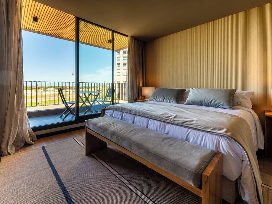 Un dormitorio con una cama grande, una banca, un balcón con vistas a edificios y un techo de madera.