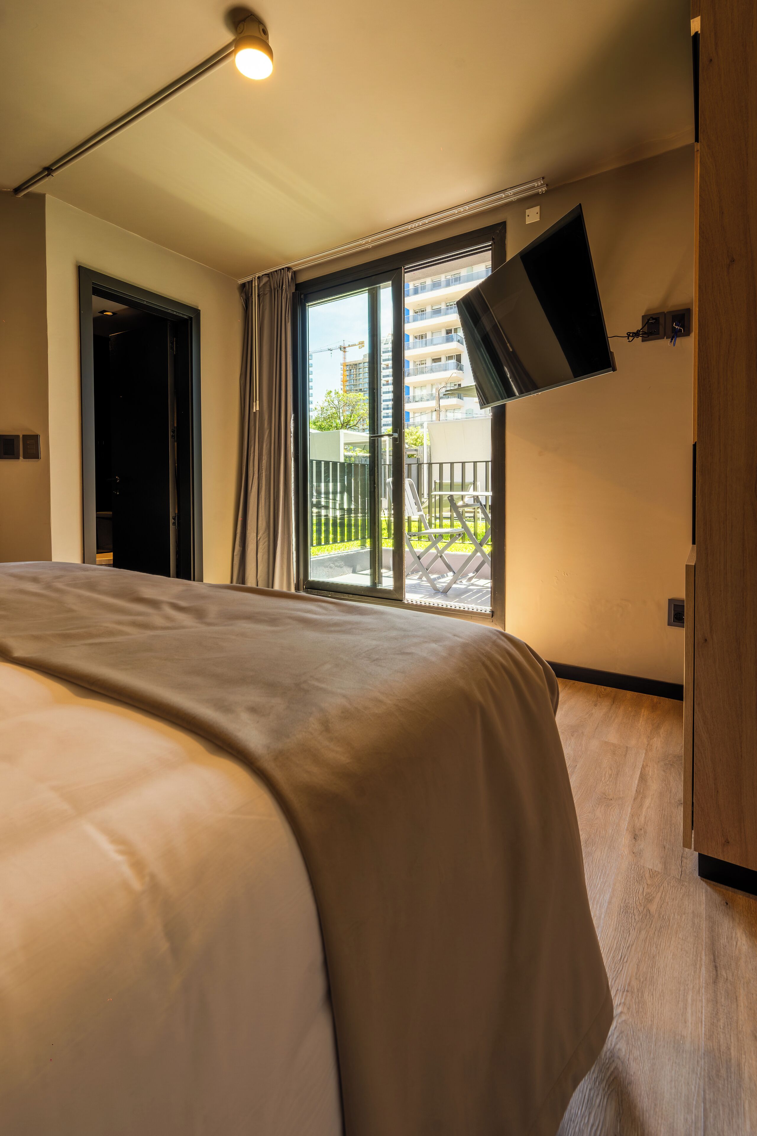 A modern hotel room with a large bed, a flat-screen TV mounted on the wall, and a sliding glass door leading to an outdoor balcony.
