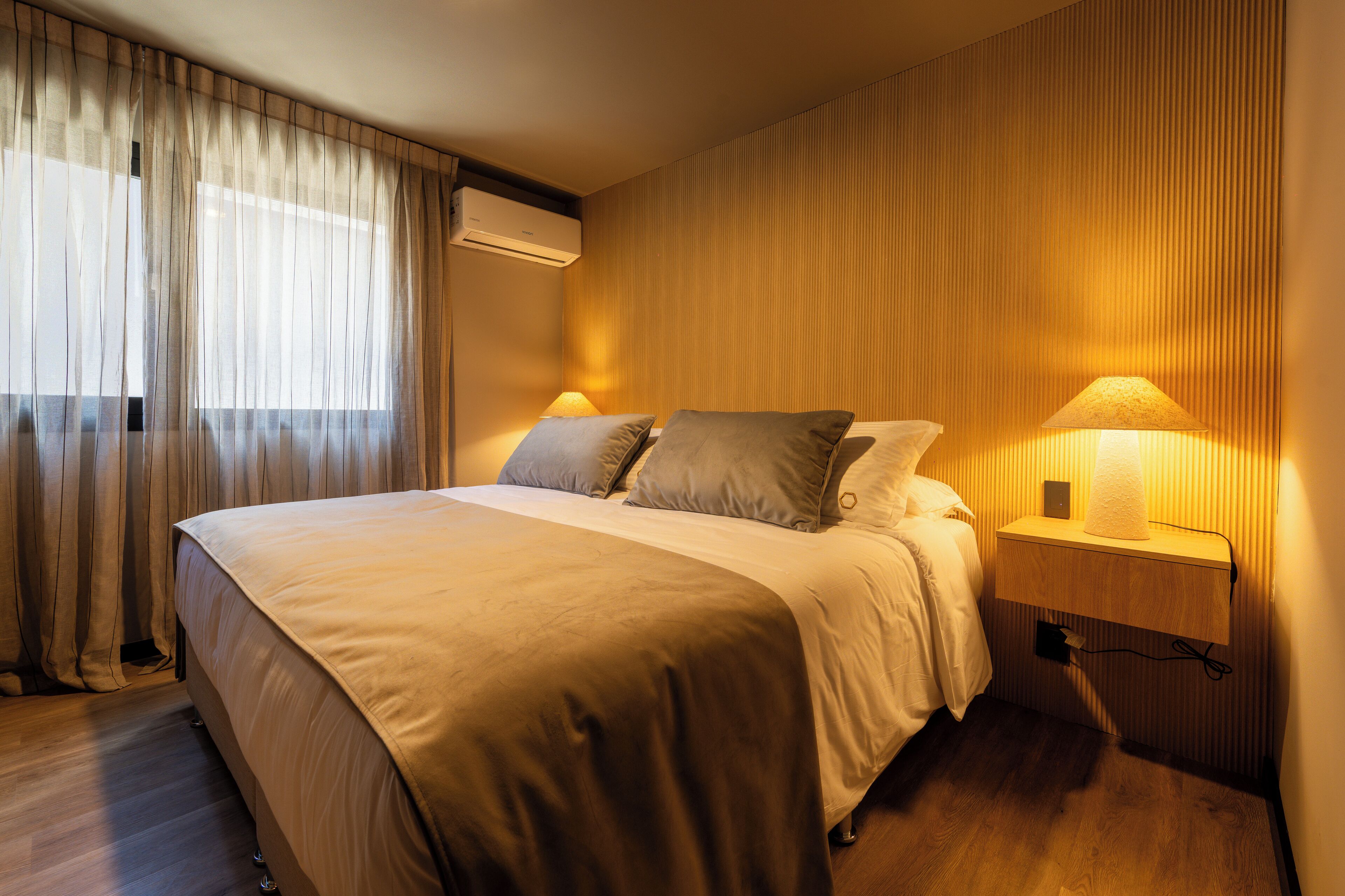 Un lit bien fait, avec une tête de lit, une table de chevet avec une lampe et un climatiseur fixé au mur.