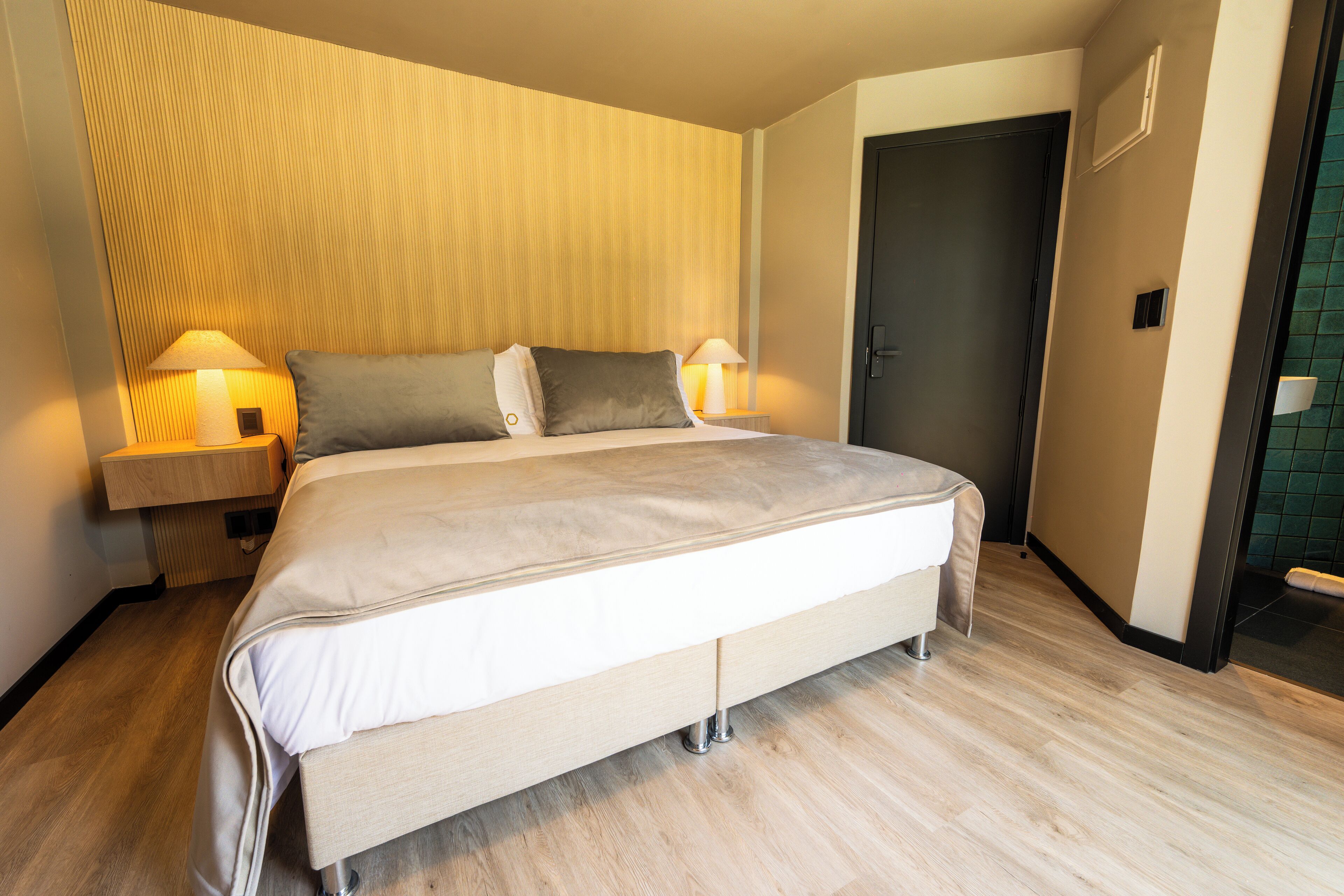 Una habitación de hotel moderna con una cama grande, dos lámparas de noche, un cabecero de madera y una puerta al baño.