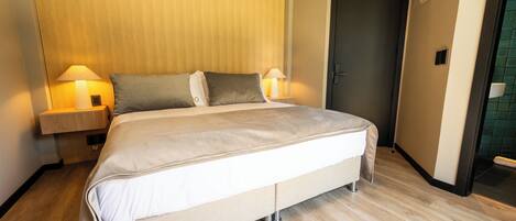 Una habitación de hotel moderna con una cama grande, dos lámparas de noche, un cabecero de madera y una puerta al baño.