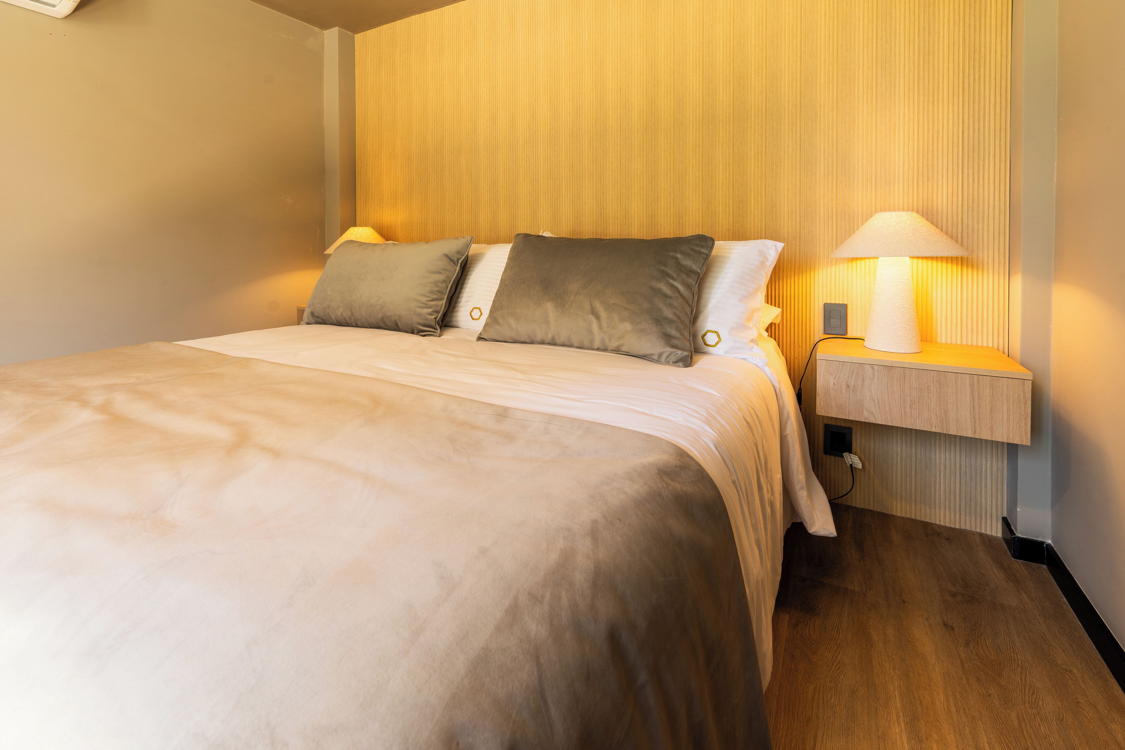 Una cama bien hecha con cabecera de madera, una mesita de noche con lámpara y un enchufe en la pared.