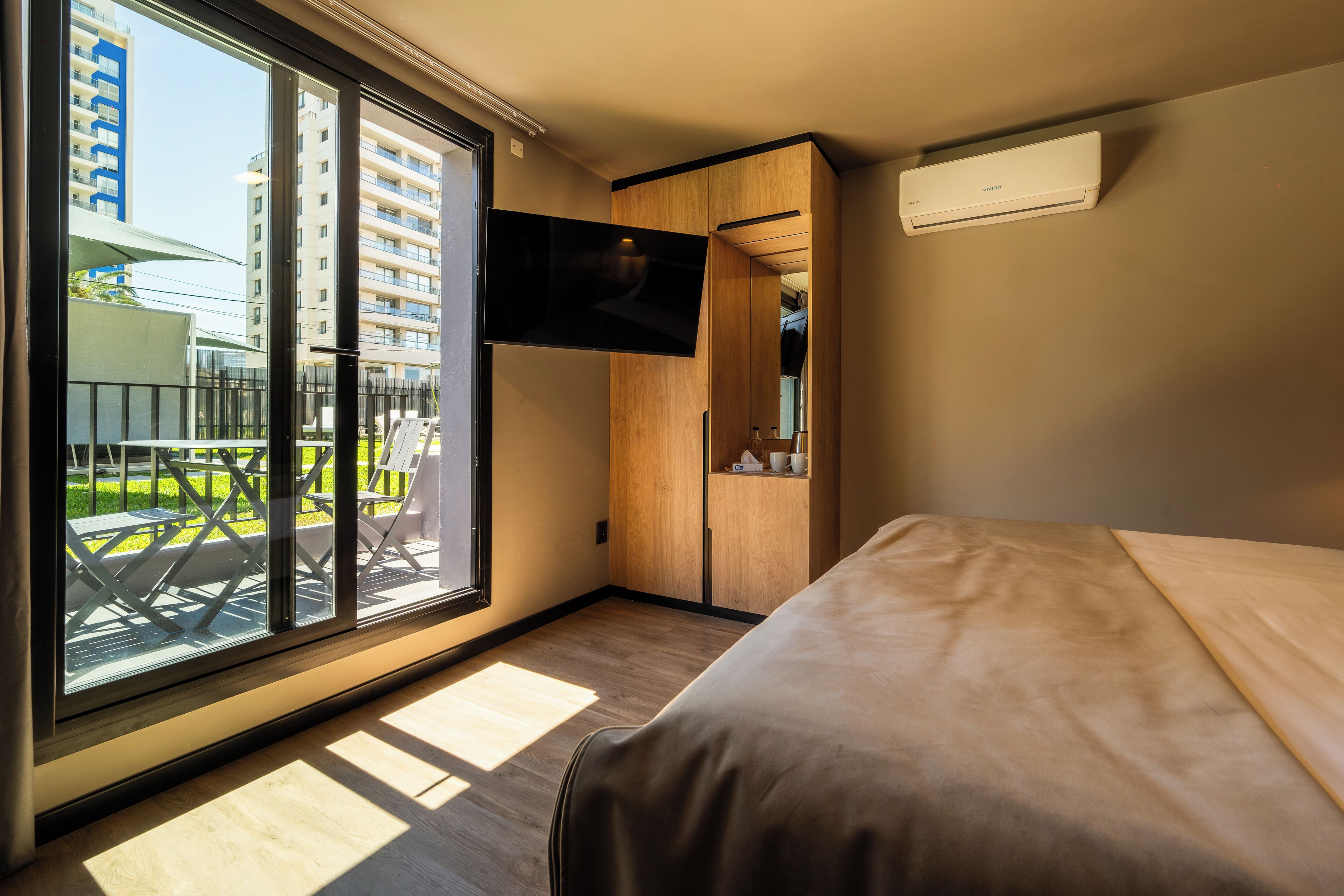 A modern hotel room with a large sliding glass door to the outside, a flat-screen TV mounted on the wall, and a bed with a wooden headboard.