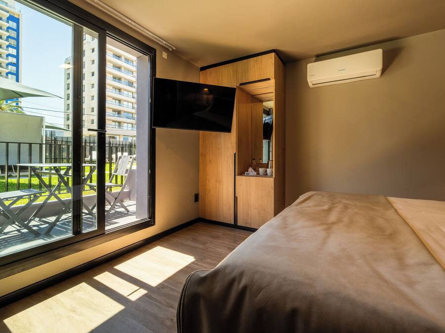 A modern hotel room with a large sliding glass door to the outside, a flat-screen TV mounted on the wall, and a bed with a wooden headboard.