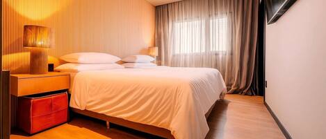Una habitación de hotel moderna con una cama grande, una mesita de noche, un mueble de almacenamiento rojo y un televisor montado en la pared.