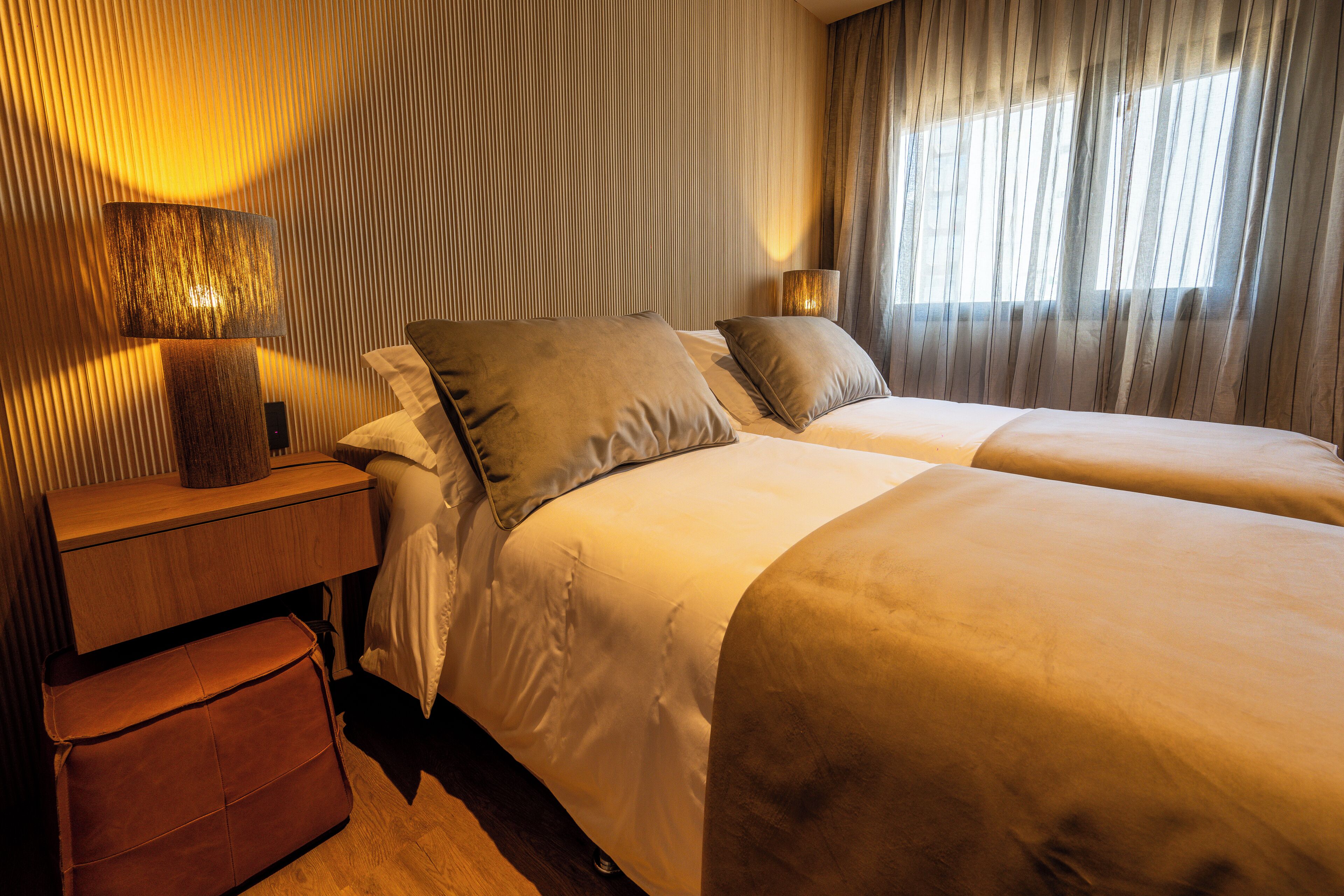 A hotel room with two beds, a wooden nightstand, a lamp, and a window with curtains.