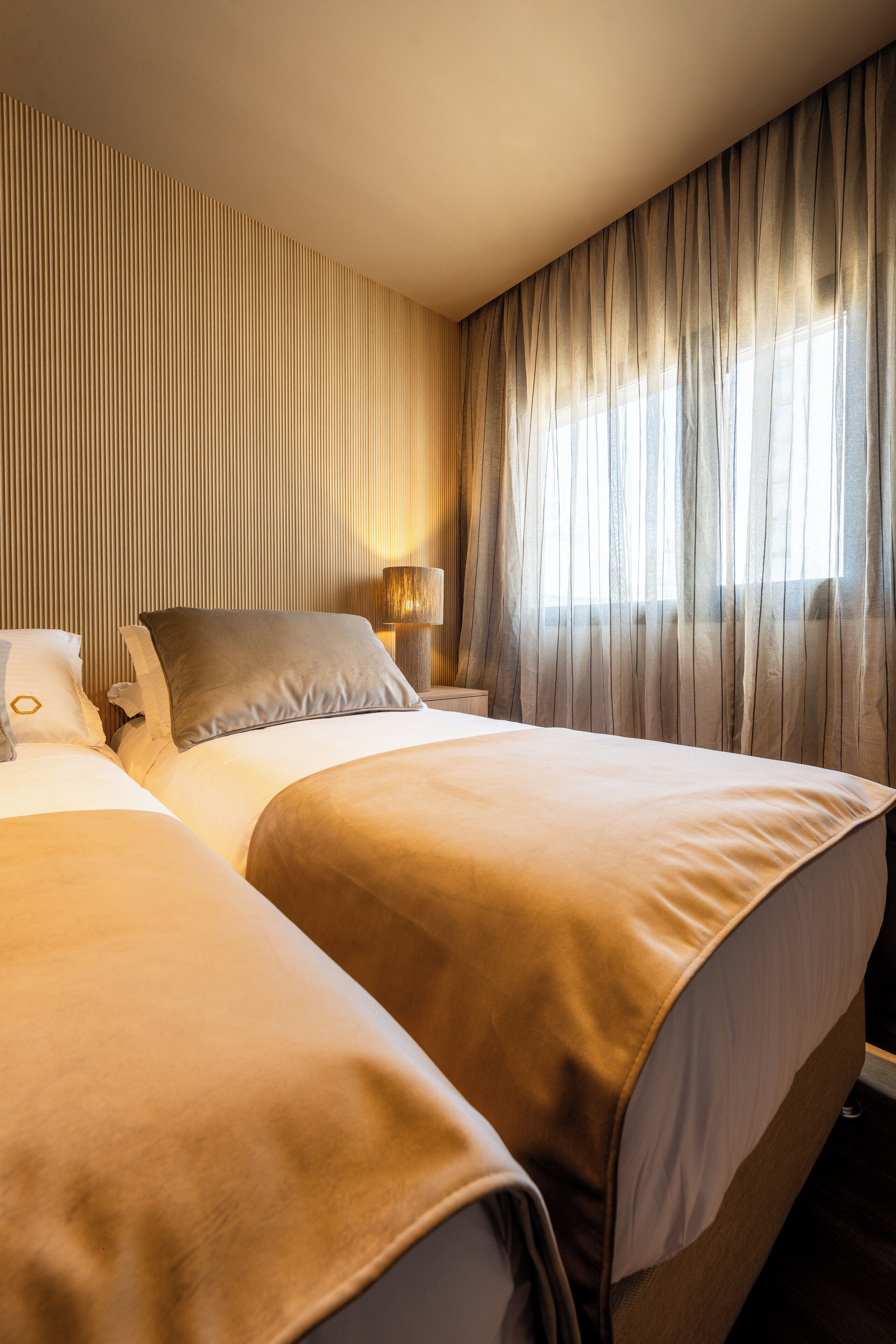 A hotel room with two beds, a nightstand, a lamp, and a large window with sheer curtains.