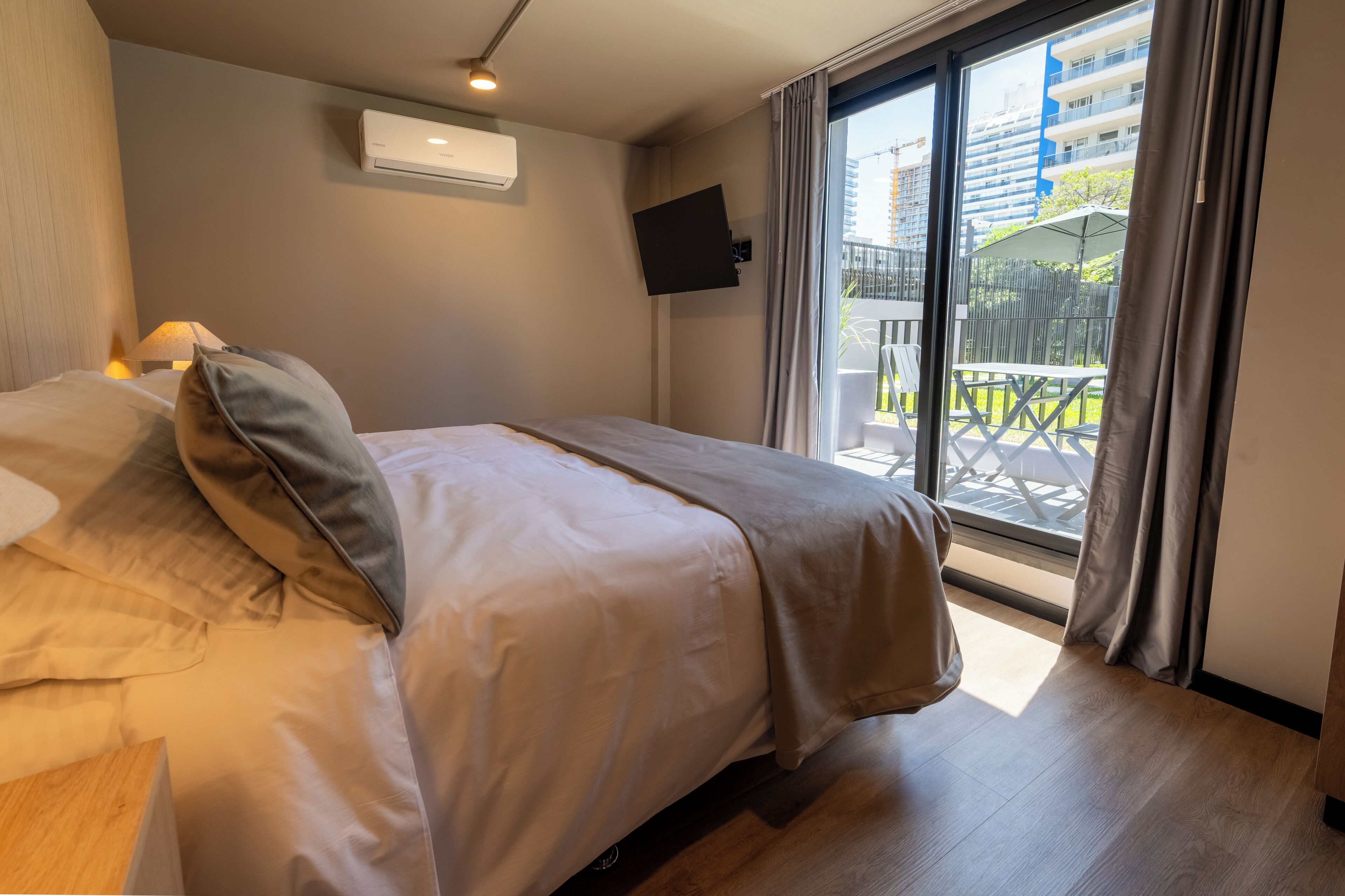 A hotel room with a bed, a TV mounted on the wall, and a balcony with outdoor furniture.