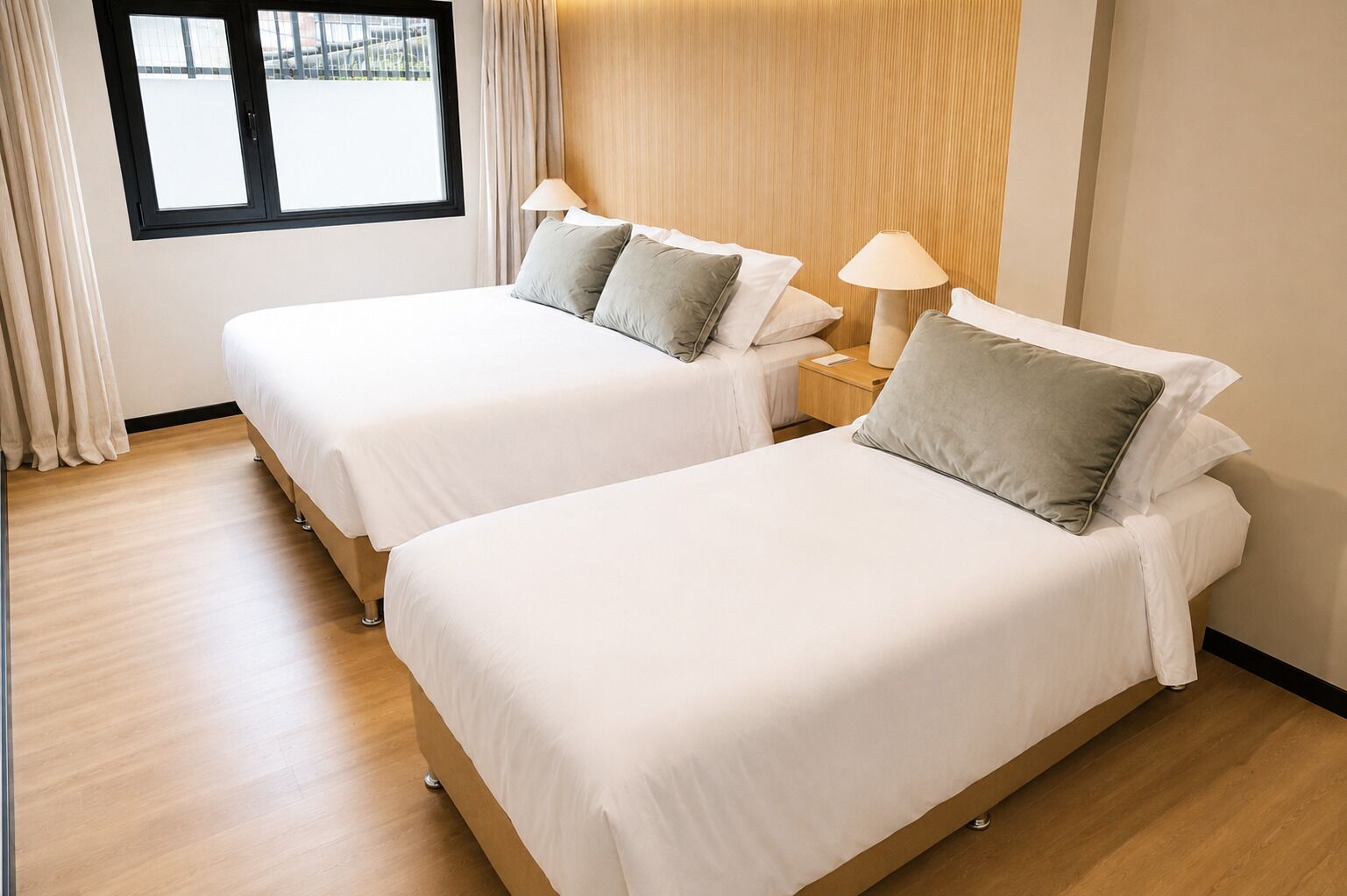 A hotel room with two single beds, a wooden headboard, a window with curtains, and a nightstand with a lamp.