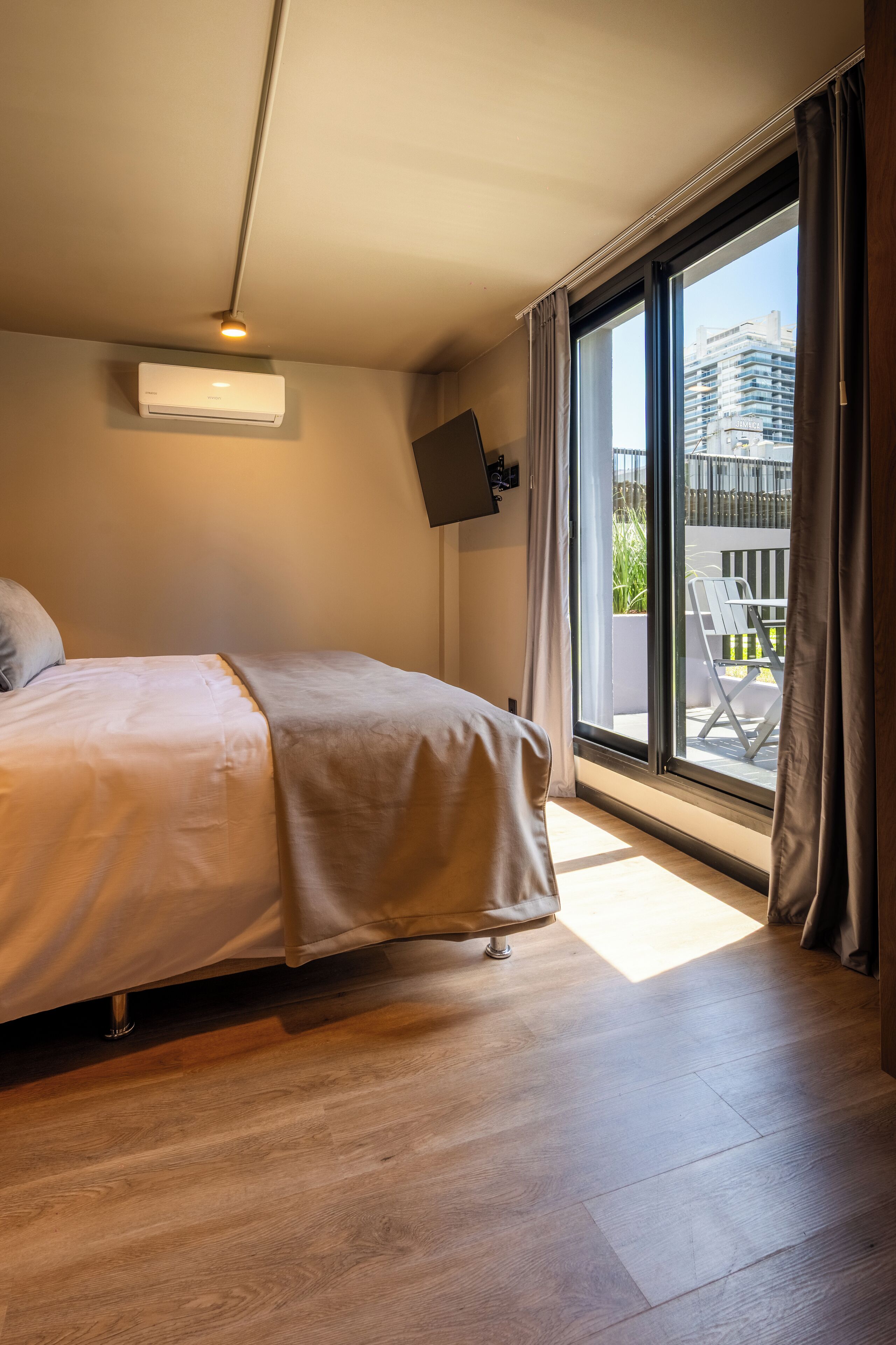 Une chambre d’hôtel moderne dotée d’un grand lit, d’un téléviseur à écran plat fixé au mur et d’un balcon avec coin salon extérieur.