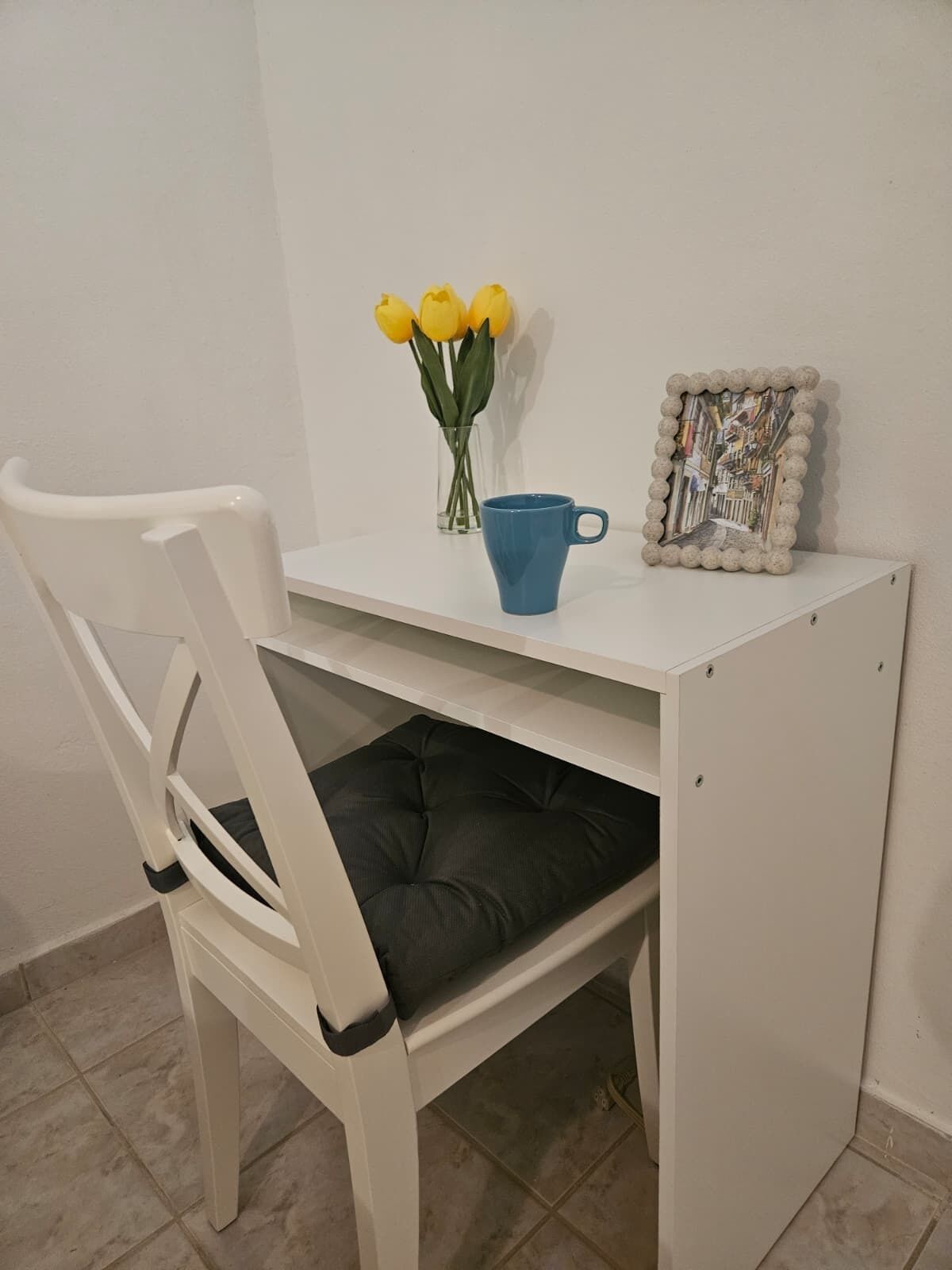 Een wit bureau met een blauwe mok, een vaas met gele bloemen en een lijst met een foto.