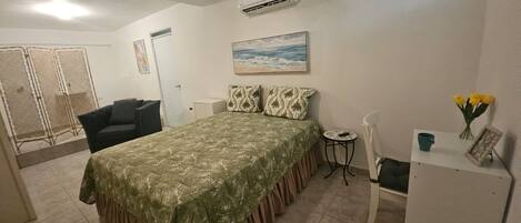 A bedroom with a bed, a chair, a small table, an air conditioning unit, a ceiling fan, a painting, and a window.