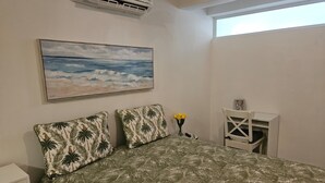 Une chambre à coucher avec un lit, un tableau représentant une plage, une petite table sur laquelle est posé un vase de fleurs et une chaise blanche.