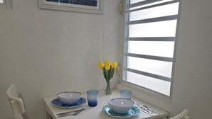 Un coin repas avec une table et des chaises blanches, un tableau représentant des bateaux accroché au mur et une fenêtre avec des stores.