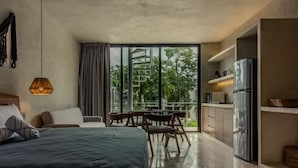 A modern bedroom with a large bed, a dining area with chairs, a kitchen with a refrigerator, and a view of greenery through large windows. - Seremonia Selva • Penthouse in the Jungle (Tulum)