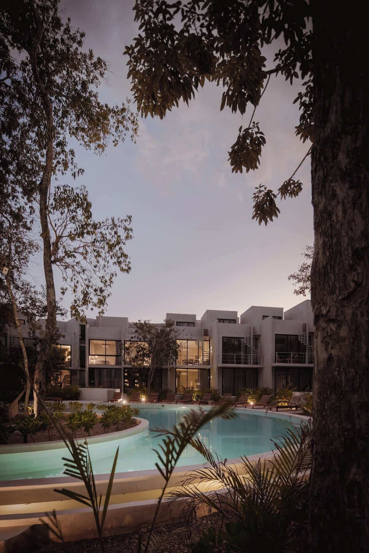 A modern hotel with a swimming pool, surrounded by trees and featuring large windows.