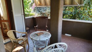 Un espacio de patio cubierto con mobiliario de mimbre, una silla de madera y vistas a zonas verdes.