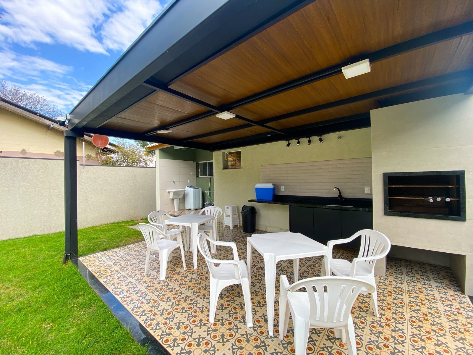 An outdoor patio with a tiled floor, white plastic chairs, a table, a barbecue grill, and a sink.