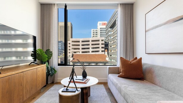 A modern living room with a sofa, coffee table, and TV on a wooden cabinet. A large window offers a view of city buildings and a parking garage.