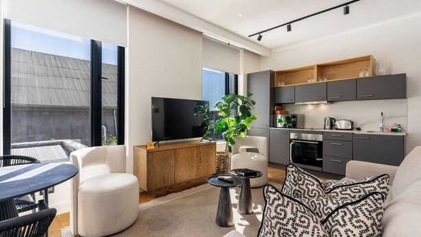 Un soggiorno moderno con un divano, un tavolino e una TV. Si vedono anche una cucina con mobili grigi e un'area pranzo con tavolo e sedie.