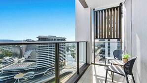 Un balcón con vistas a un paisaje urbano moderno, barandilla de cristal y una mesa con sillas.