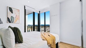 Un dormitorio con una cama grande, vistas de la ciudad a través de ventanales del suelo al techo y un cuadro en la pared.
