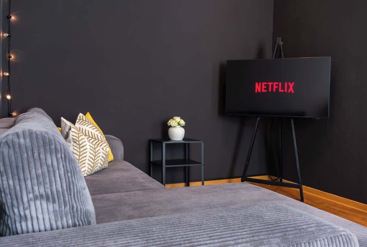 Moderni olohuone, jossa on harmaa sohva, pieni musta sivupöytä, jossa on maljakko, ja suuri, litteänäyttöinen televisio, jonka mustalla jalustalla näkyy teksti ’NETFLIX’.