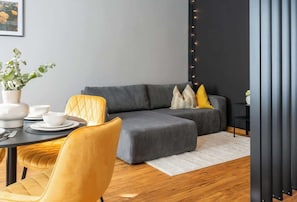 Ein modernes Wohnzimmer mit einem grauen Ecksofa, gelben Sessel, einem Esstisch mit weißen Tellern und Schüsseln sowie einer Topfpflanze.