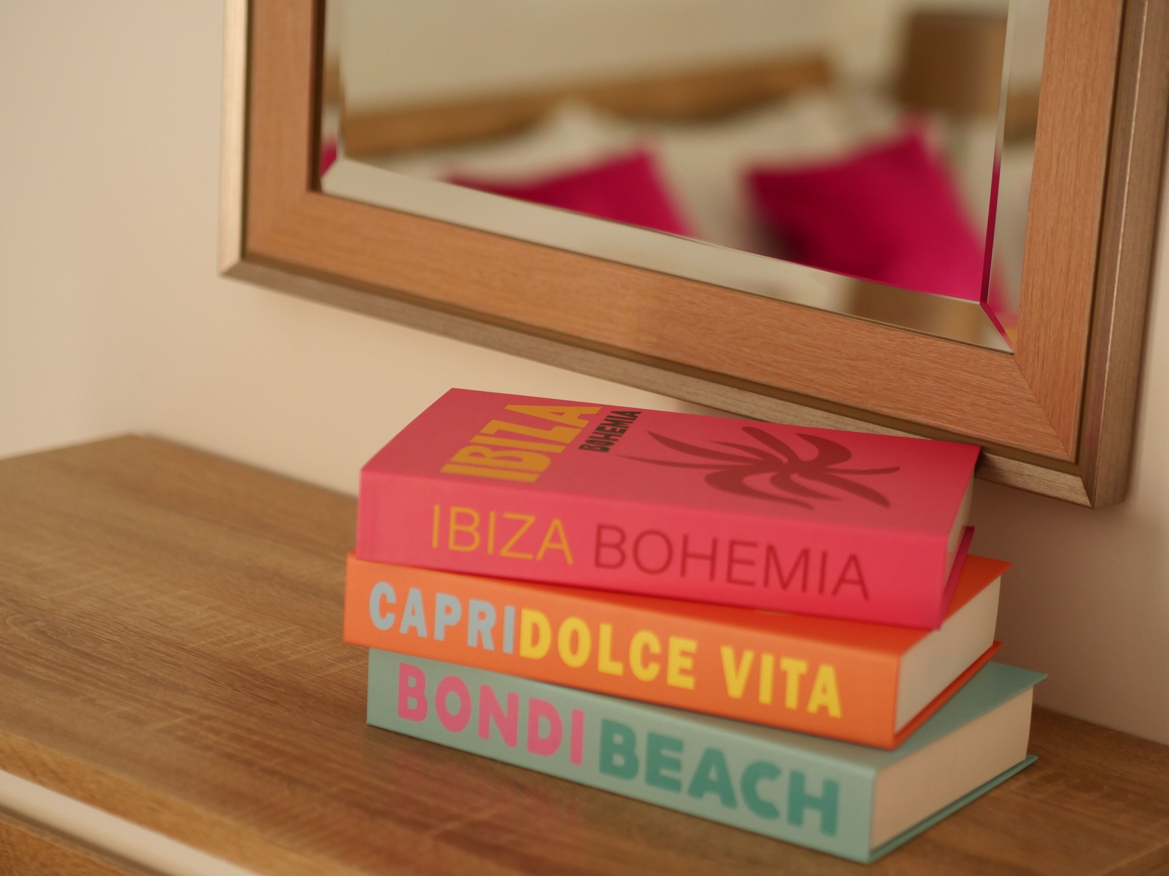 A stack of books on a wooden surface with a mirror reflecting a pink pillow.