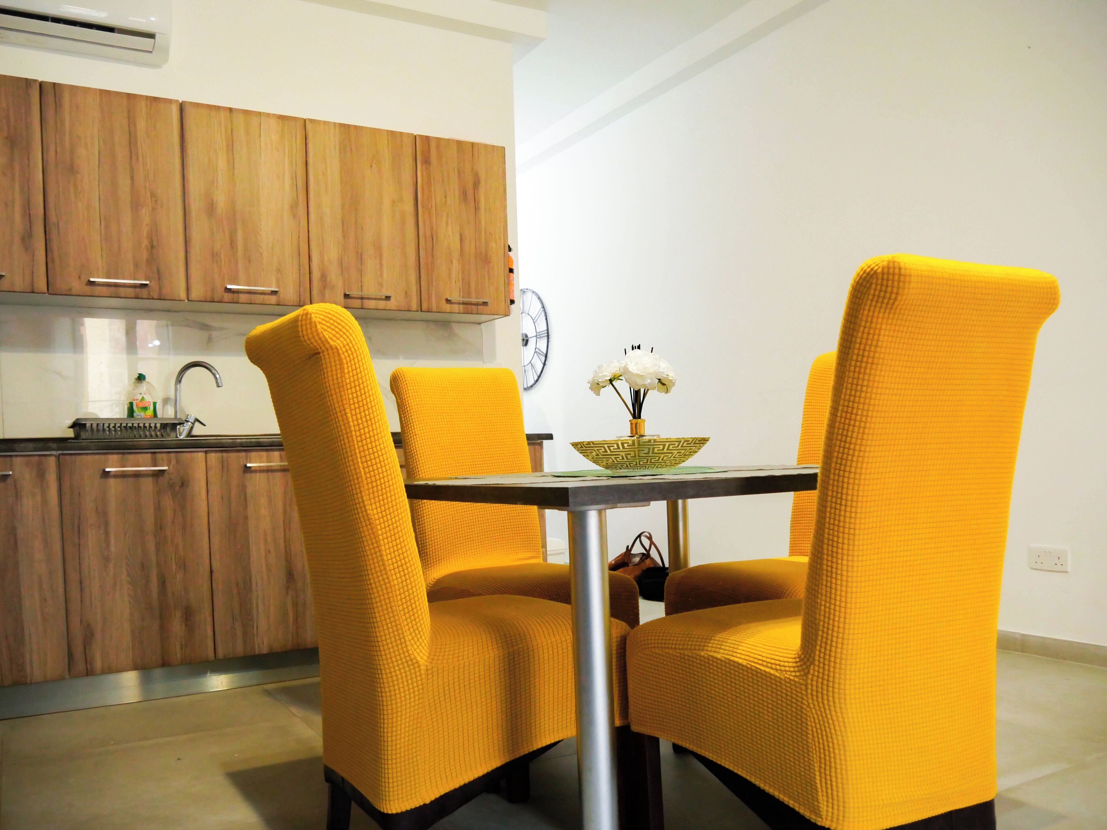 Un comedor con una mesa y cuatro sillas amarillas, un reloj en la pared y armarios de madera al fondo.