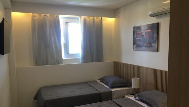 Una camera d'albergo piccola con due letti singoli, una finestra con tende, un condizionatore d'aria a muro e un quadro raffigurante autobus.