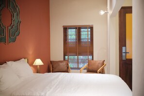 A hotel room with a bed, two chairs, a window with blinds, a lamp, and a door.