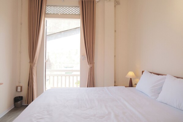 Habitación de hotel con cama, cortinas, mesita de noche con lámpara y vista al balcón exterior.