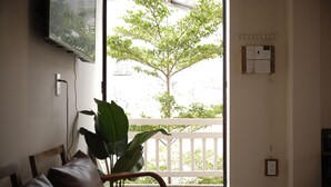 A room with a wooden chair, a large potted plant, a television mounted on the wall, and a view of a balcony with a tree. - DAINOI Hue Homestay (Hue)