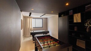 A room with a foosball table, a window with curtains, and a bookshelf with plants. - Apto em Pinheiros c Piscina Academia e Smartlock (São Paulo)