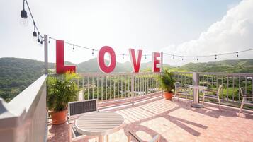 Varanda com vista para colinas verdes, um letreiro com a palavra 'LOVE', luzes de Natal e móveis de jardim.