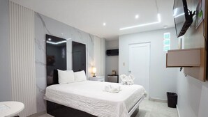 A modern hotel room with a large bed, a bedside table, a TV mounted on the wall, and a small desk with a chair. - Hotel Vista Verde (Yauco)