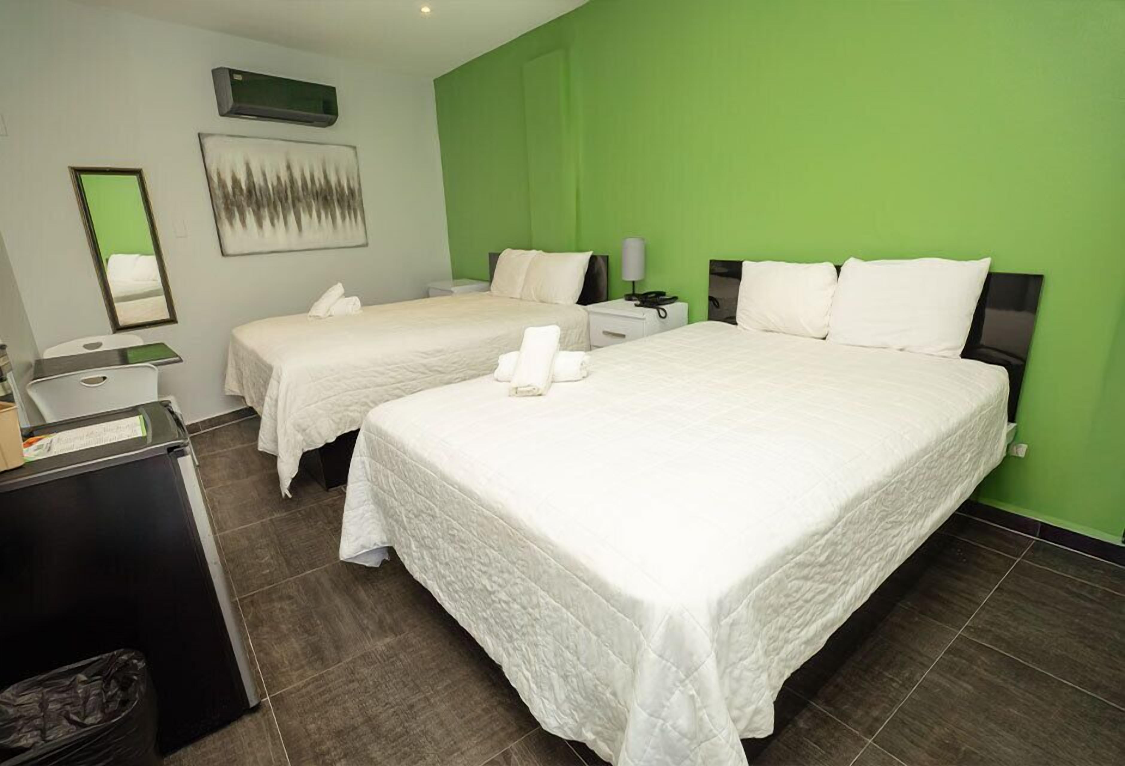 A hotel room with two beds, a desk, a trash bin, and a green accent wall.