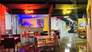 A brightly lit indoor dining area with tables and chairs, neon signs, and a vending machine. - Hotel Vista Verde (Yauco)