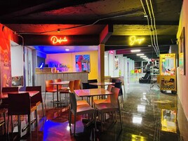 Un área de comedor interior luminosa con mesas y sillas, letreros de neón y una máquina expendedora.