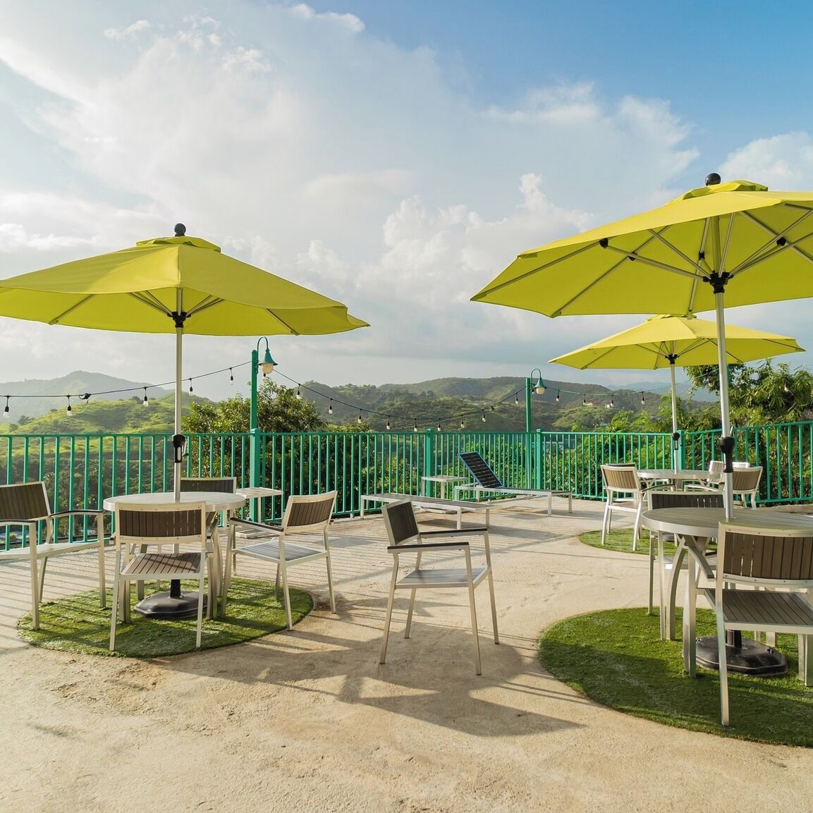 An outdoor patio with yellow umbrellas, white tables, and chairs, surrounded by a green fence and overlooking a scenic landscape.