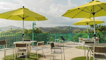 An outdoor patio with yellow umbrellas, white tables, and chairs, surrounded by a green fence and overlooking a scenic landscape.