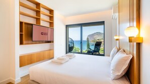 A hotel room with a large bed, a TV on the wall, a balcony with a chair, and a view of the sea.