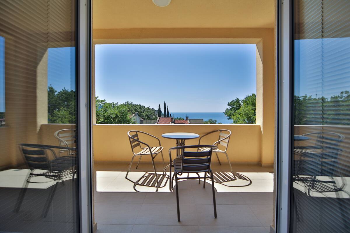 A balcony with a table and chairs, offering a view of the sea and greenery.