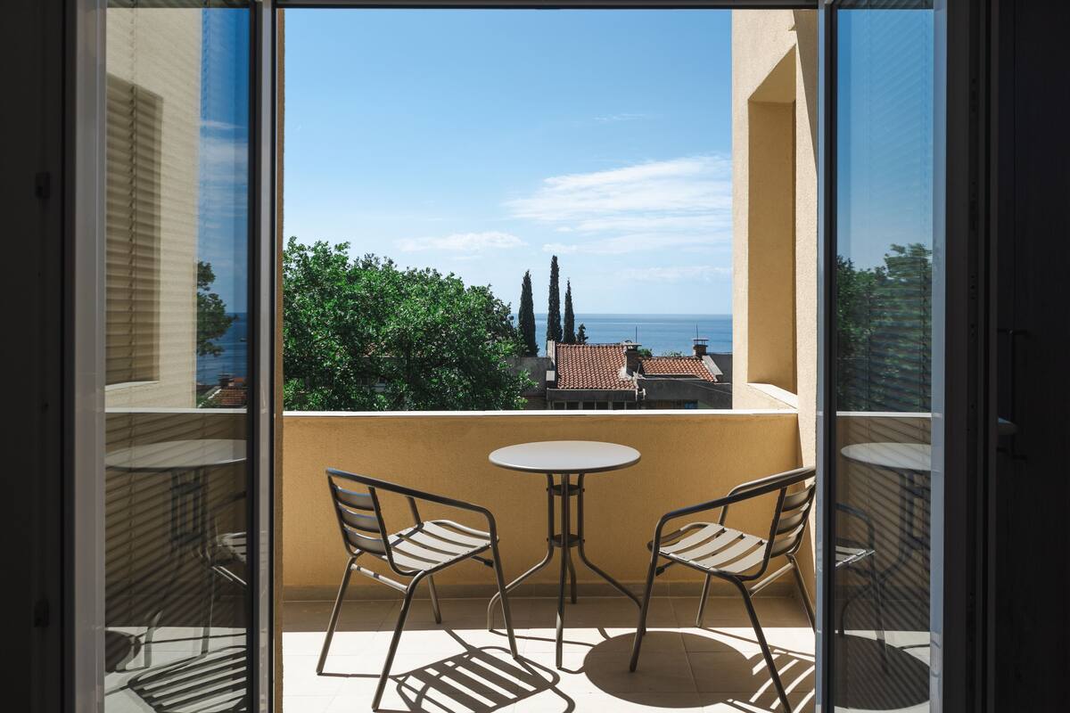 A balcony with two chairs and a table, overlooking a coastal view with trees and buildings.