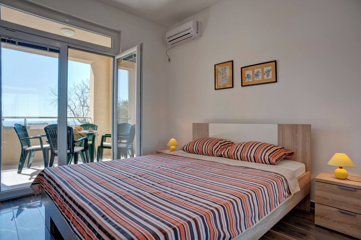 A bedroom with a large bed, a nightstand with a lamp, and a sliding glass door leading to an outdoor area with chairs.
